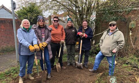 Group of men and women stand on patch of soil with big forks and spades