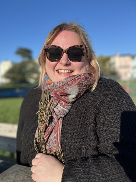 Head and shoulders shot of Carrie sat at bench, in black jumper with pink scarf and sunglasses