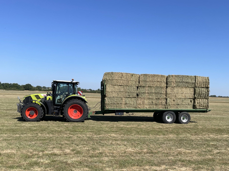 Green tractor carrying a trailer of hay bales across field, with clear blue sky in background